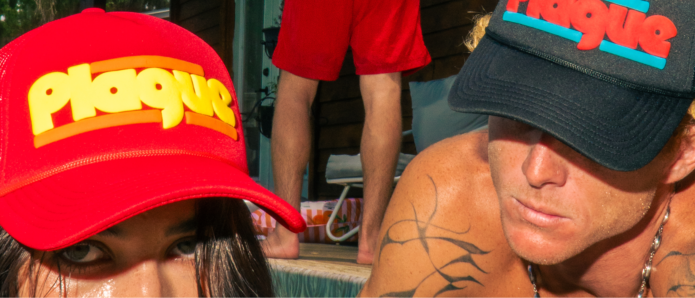 Two people wearing Plague brand trucker hats in an outdoor setting. One person in the foreground wears a bright red hat with yellow and orange lettering, while the other wears a black hat with red and blue lettering.